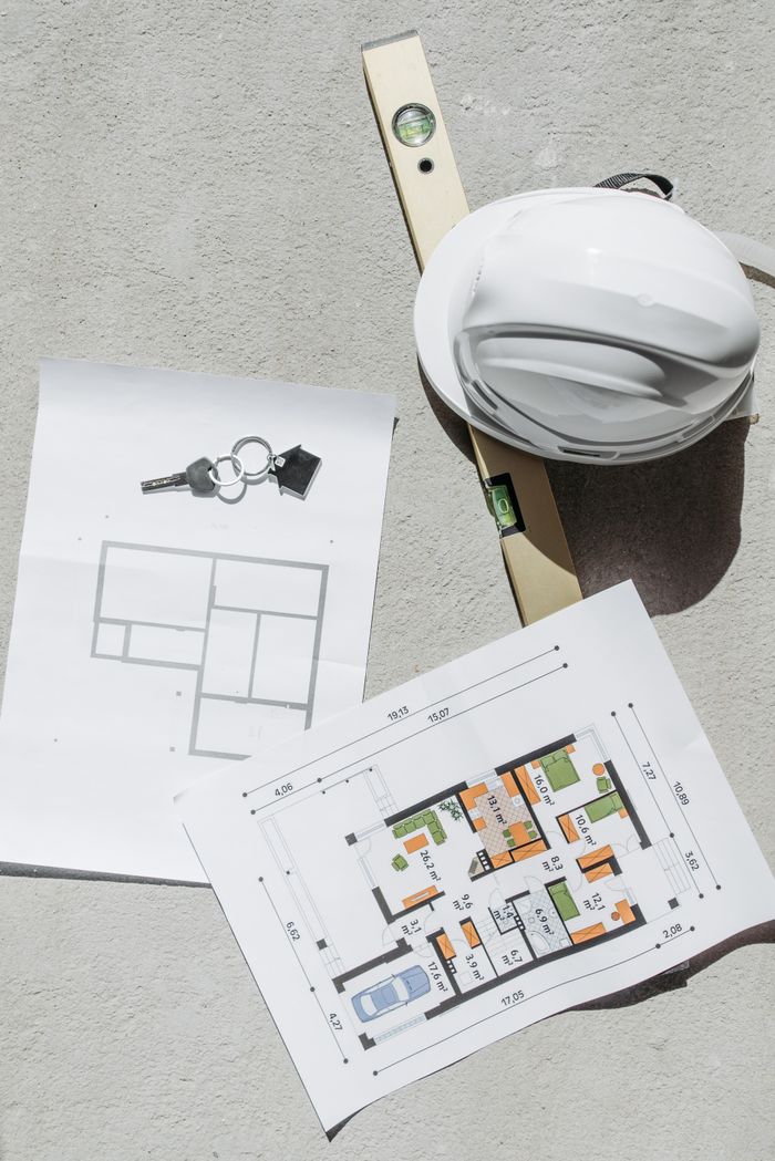 gallery-6 Flat lay of construction plans, keys, hard hat, and level tool on concrete.