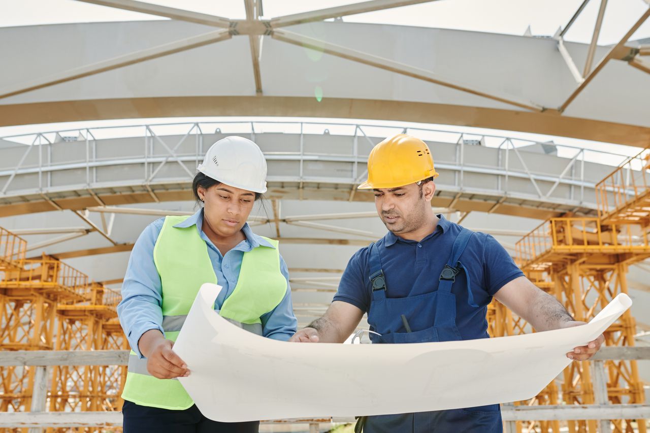 services-03 Male and female engineers discussing blueprints at a construction site, promoting teamwork and planning.